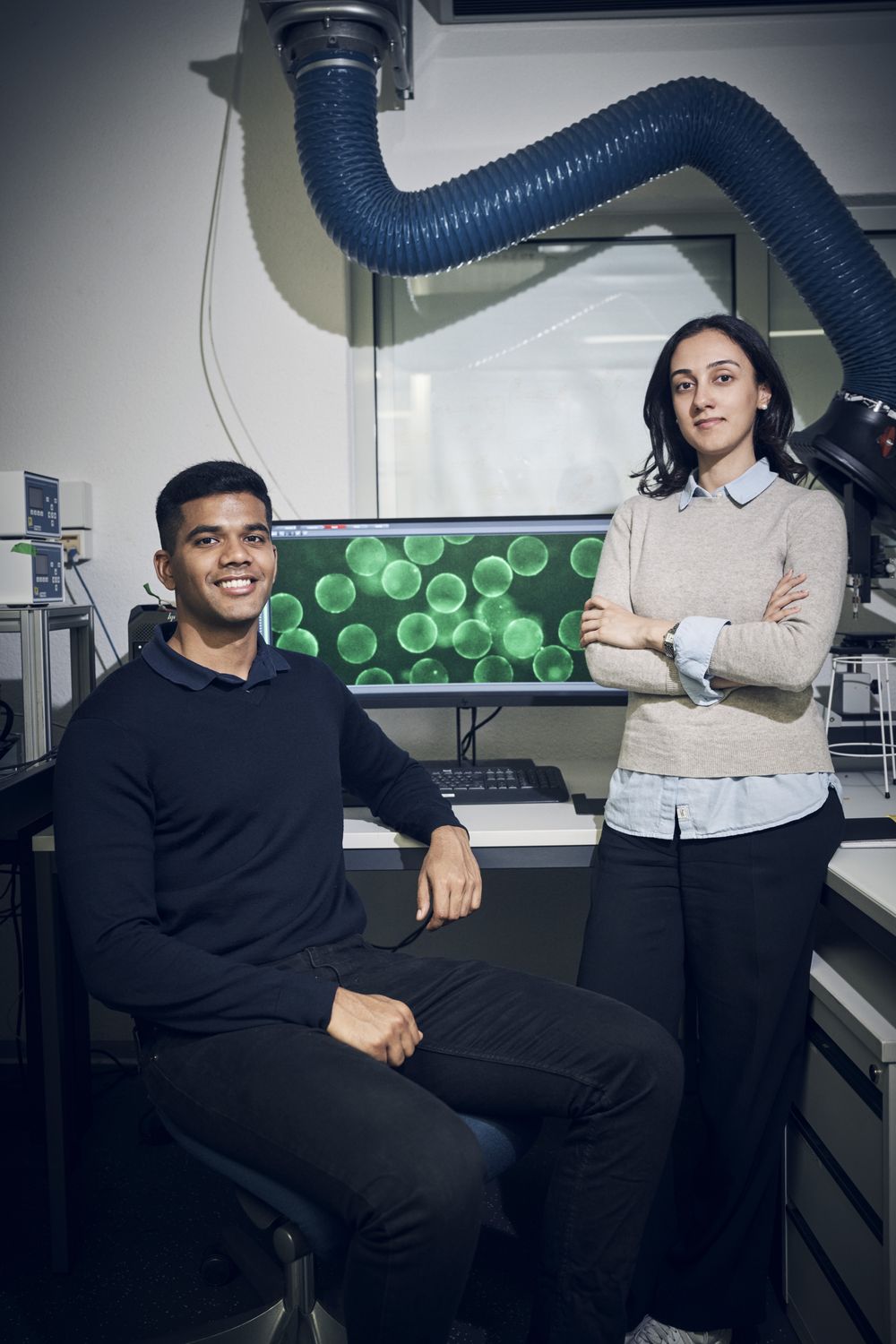 Apoorv Singh, seated, and Börte Emiroglu, standing, in the lab in front of a screen showing green fluorescent microgels
